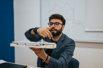 A teacher gesturing as he explains an architectural model in a classroom. Engaged in teaching, he...