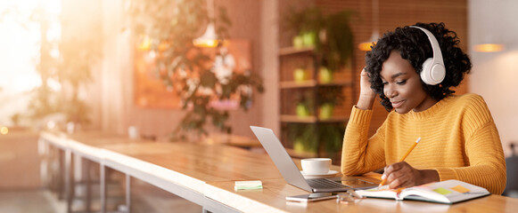 Smiling black girl with wireless headset studying online, using laptop at cafe, taking notes, copy...