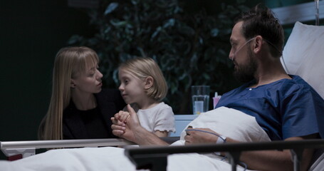 Sick husband in hospital bed wife and daughter sit beside sick man while nurse comes and father signs concent agreement.