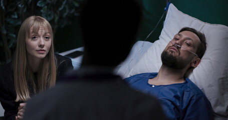 Man in hospital bed, girlfriend sitting beside, talking and supporting.