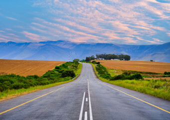 Road rolls over hills covered in golden wheat fields, with mountains in the distance