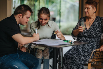 A boy with Down syndrome assists a girl in a wheelchair with an art project, while a woman supports...
