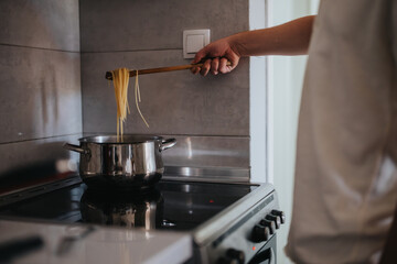 A person's hand using a wooden spoon to cook spaghetti on a stove in a contemporary kitchen setting, capturing a home cooking moment.