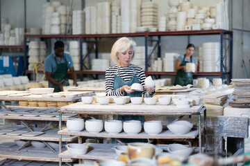 Mature female craftswoman checking quality of pottery in pottery factory