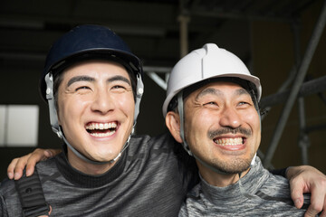 現場作業員(鳶職や屋外の安全帯ハーネスをつけた）の肩を組むガテン系仲良し2人の見上げる笑顔　転職や採用のイメージのクローズアップ