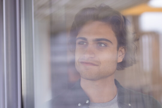 Young man gazing thoughtfully through window, reflecting on holiday moments, at home