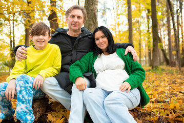 Fototapeta premium Happy family in autumn park. Father, pregnant mother and son sitting on fallen leaves. 