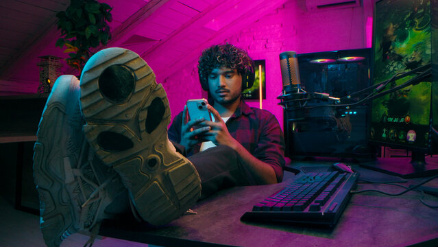 Indian American man gamer gambler computer game technology mobile phone smartphone pc addiction pause network keyboard excited male mouse put feet table gadgets guy stuck connection cyber electronic