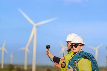 Two engineers wearing high visibility safety jackets and helmets measure wind speed with an...