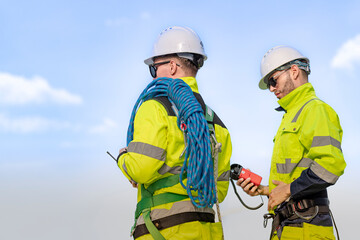 Two engineers in high visibility jackets and helmets are inspecting a wind farm. One is holding a mobile device, while the other is equipped with ropes for climbing, with turbines visible .