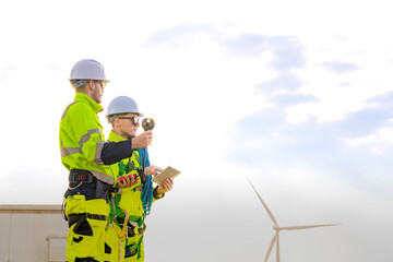 Two engineers wearing high visibility safety jackets and helmets measure wind speed with an...