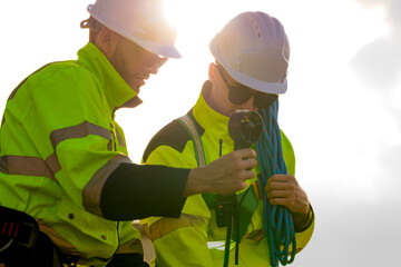 Two engineers wearing high visibility safety jackets and helmets measure wind speed with an...