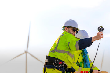 Two engineers wearing high visibility safety jackets and helmets measure wind speed with an...