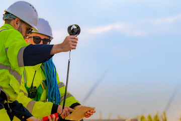 Two engineers wearing high visibility safety jackets and helmets measure wind speed with an...