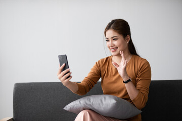 Naklejka premium Young Woman Sitting and Relaxing on a Sofa, Smiling and Waving at Smartphone, Casual and Comfortable Home Setting, Modern Lifestyle, Indoor Relaxation, Happy and Connected, Technology and
