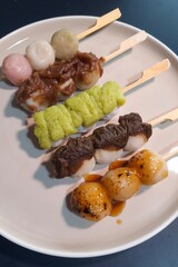 Mitarashi, Hanami, Houjicha, Zunda, and Yomogi anko dango on a white plate on navy table. Dango is a traditional sweet in Japan.