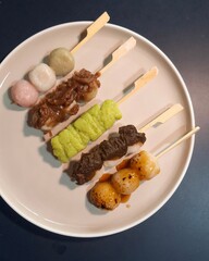 Top view of Mitarashi, Hanami, Houjicha, Zunda, and Yomogi anko dango on a white plate on navy table. Dango is a traditional sweet in Japan.