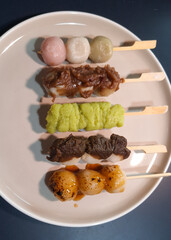 Top view of Mitarashi, Hanami, Houjicha, Zunda, and Yomogi anko dango on a white plate on navy table. Dango is a traditional sweet in Japan.