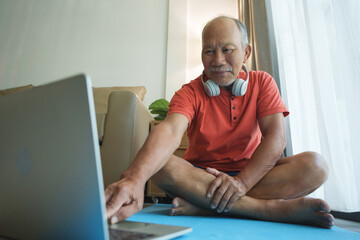 Asian elderly man wearing headphones is sitting on a yoga mat in front of his laptop.