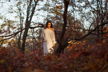 A young brunette woman in a cozy white off-shoulder sweater walks through a peaceful autumn forest, bathed in soft natural light. Ideal for fall fashion, seasonal lifestyle, and nature-related