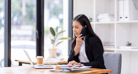 Obraz premium asian woman talking on the phone while working on computer in desk office. Searching on internet 