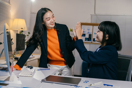 Two young businesswomen high-fiving in office at night, celebrating success of new project together. Sitting at desk with laptops, working as team - Powered by Adobe