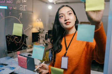 Young businesswoman is using adhesive notes to plan a project on a glass wall in an office. She is...