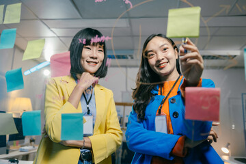 Two asian businesswomen are using colorful sticky notes and a marker pen, working together on a project, brainstorming and planning strategy on a glass wall in the office at night