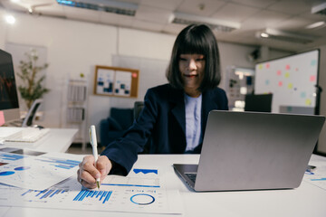 Focused businesswoman analyzing graphs and taking notes at office desk with laptop, delving into finance, accounting, strategy, planning, marketing, and sales data