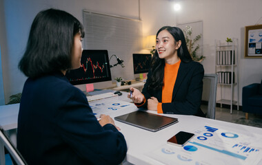 Two businesswomen are having a productive meeting in the office, analyzing charts and graphs displayed on their computer screen and printed documents