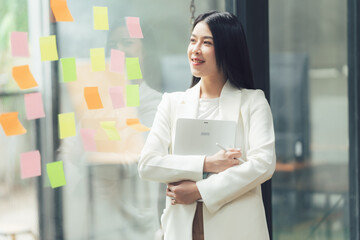 Confident Businesswoman Planning: A young, stylish businesswoman stands confidently by a window, holding a tablet, surrounded by colorful sticky notes, radiating ambition and success. 