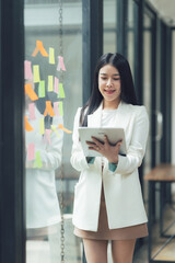 Focused Businesswoman Planning Strategy: A young, smiling Asian businesswoman uses a tablet, reviewing colorful sticky notes on a glass wall, showcasing her strategic planning and innovative approach.