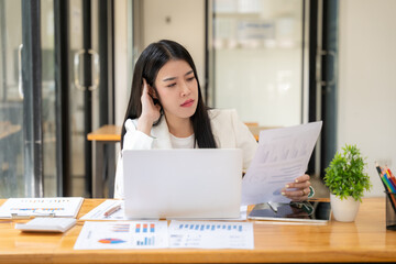 Focused Businesswoman Reviewing Documents: A young, serious Asian businesswoman meticulously examines financial documents at her desk, laptop and tablet nearby.