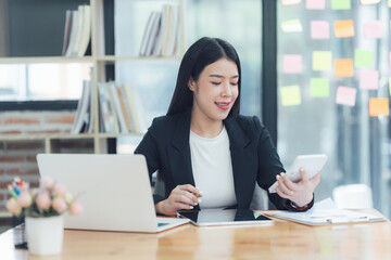 Fototapeta premium Focused and Determined: A young businesswoman exudes confidence as she works at her desk, her eyes fixed on her phone, showcasing a dedication to her craft. The laptop, pen.