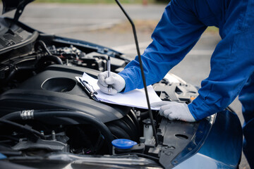 Hand of a professional Asian mechanic in blue workwear, writing on a paper while repairing a car in an auto repair shop. Close-up shot.
