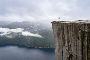 A woman stands at the edge of Preikestolen cliff, overlooking the vast Lysefjord landscape,...