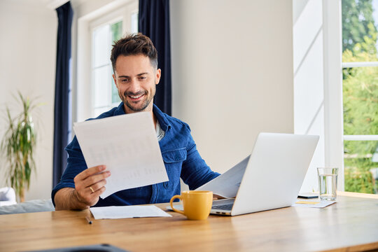 Happy man looking at financial documents at home sitting at a table with laptop in living room