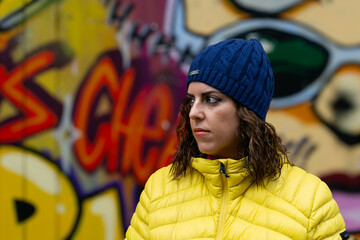 A brunette woman, in her 30s, wearing a yellow puffer jacket and a blue wool hat, looking serious...