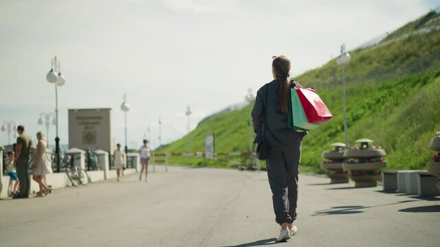 Woman in grey clothing walks with shopping bags over her shoulder, people stand by the bridge's edge with parked bicycles, a roadblock and flower vest are visible on the sunny day