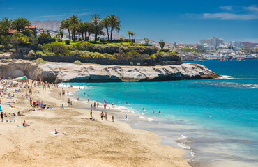 Playa del Duque offers a serene beach day with golden sand, clear blue water, and a bustling crowd enjoying the sun
