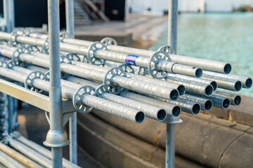 Several galvanized metal scaffolding pipes are stacked and secured on a frame at a bustling construction site next to a serene waterfront.