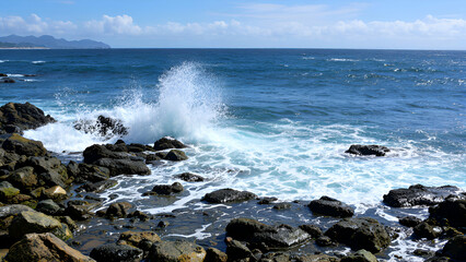 A wave crashes on a rocky shore, creating a spray of white foam. The blue ocean extends to the horizon, with a distant mountain range visible.