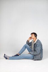 Chico joven vestido de jeans con auriculares, escuchando música o podcast, sentado en el suelo...