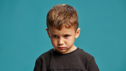 Little boy stares intently with a serious, determined expression against blue studio background. Concept of facial-expressions, positive and negative emotions. Ad