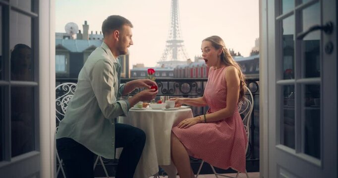 Romantic Proposal Unfolds on a Parisian Balcony as a Young Man Kneels with a Ring to Ask His Girlfriend to Marry Him. Eiffel Tower in the Background. Her Surprise Turns to Joy as She Accepts