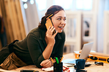 Laptop, phone call and smile of carpenter woman in workshop for professional joinery or woodworking. Computer, craftsmanship and creative with happy artisan in industrial warehouse for engineering