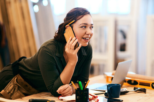 Laptop, phone call and smile of carpenter woman in workshop for professional joinery or woodworking. Computer, craftsmanship and creative with happy artisan in industrial warehouse for engineering