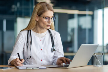 Mature female doctor working on laptop in modern office. Wearing stethoscope, taking notes, conducting research effectively. Emphasizes professionalism, focus, dedication in healthcare setting.