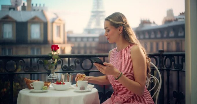 Beautiful Stylish Female Sitting Comfortably on a Balcony in Paris, Enjoying Coffee and Pastry while Scrolling Through Her Smartphone. Young Woman Online Shopping on a Phone App and Checking Updates