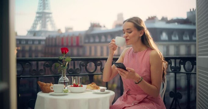 Portrait of a Young Woman Enjoying a Quiet Morning on a Parisian Balcony, Sipping Coffee and Browsing Her Smartphone to Stay Connected. Eiffel Tower Stands Majestically in the Background